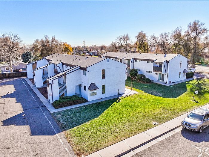Charming apartment complex with white exteriors, lush green lawns, and ample parking in a serene neighborhood setting.