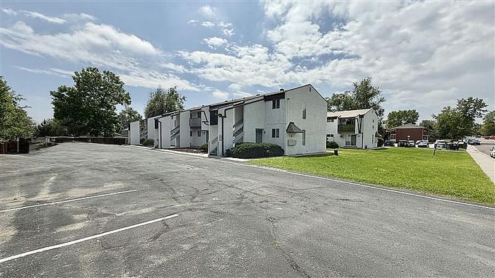 Charming white apartment complex with lush greenery and ample parking under a bright, blue sky.