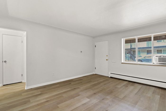 Bright and airy living space with sleek wood flooring, large window for natural light, and access to a cozy room.