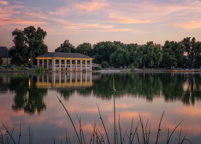 Serene lakeside pavilion with glowing lights reflecting on tranquil water at sunset, surrounded by lush greenery.