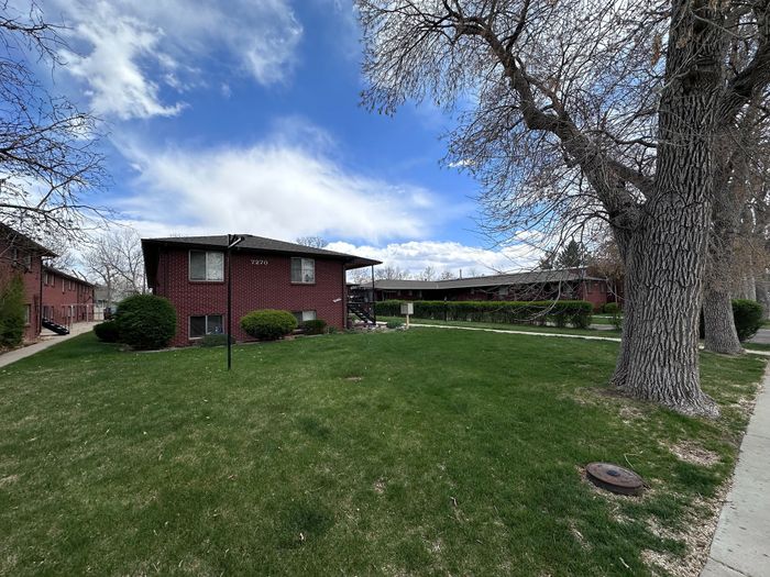 Charming brick apartment building nestled in a lush green yard with mature trees and a vibrant blue sky backdrop.