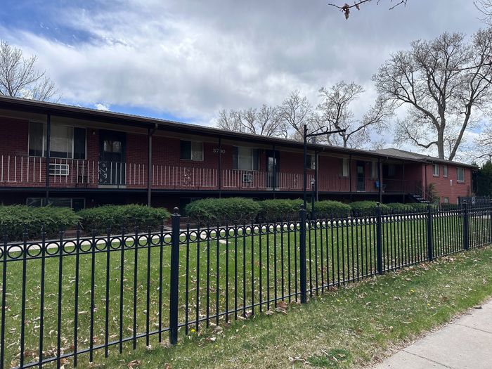 Charming brick apartment building with spacious green lawn and elegant wrought-iron fence, offering serene living in a picturesque neighborhood.