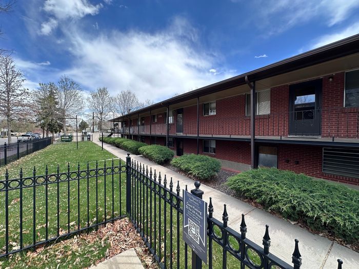 Charming brick apartment building with inviting balconies, manicured landscaping, and a welcoming sidewalk entrance under a bright, blue sky.