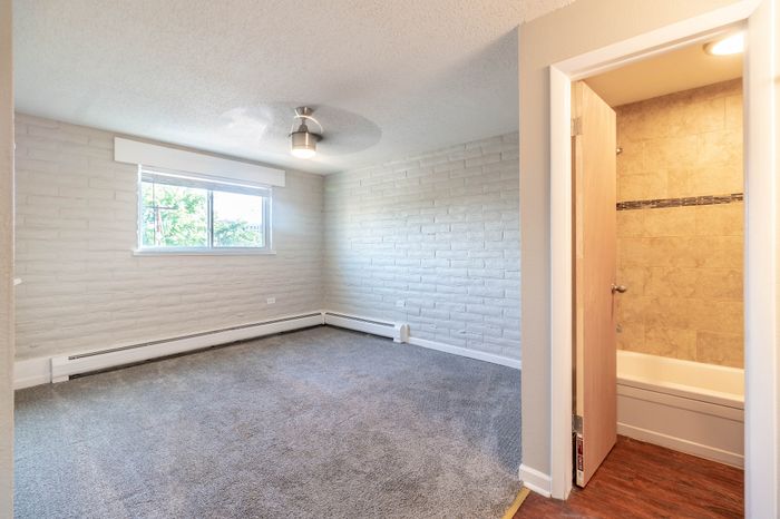 Bright and inviting bedroom with textured brick walls, cozy carpeting, and a window view of greenery, adjacent to a modern tiled bathroom.