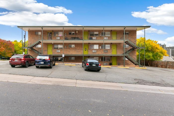 Charming two-story apartment building with vibrant green doors, ample parking, and a picturesque backdrop of autumn foliage.