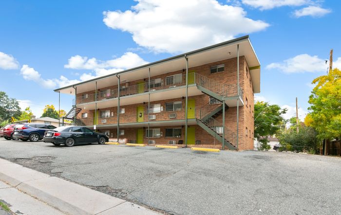 Charming brick apartment building with vibrant green doors, surrounded by lush trees and ample parking, under a clear blue sky.