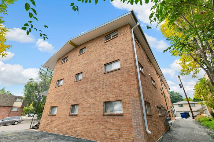 Charming brick apartment building with a vibrant exterior, surrounded by lush trees and a clear blue sky, offering a serene and inviting living environment.