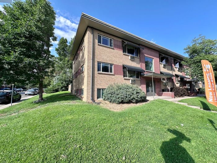 Charming two-story brick apartment building with lush green lawns, large windows, and a welcoming entrance. Now leasing sign signifies immediate availability in this appealing neighborhood.