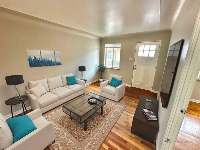 Inviting living room with elegant white sofas adorned with vibrant teal cushions, complemented by sleek black lamps and a sophisticated glass coffee table on a classic rug. Sunlit ambiance highlights the polished wooden floors and contemporary decor.