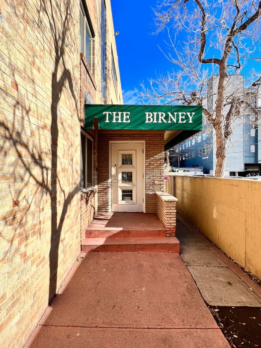 Inviting entrance of The Birney apartment building with a charming brick facade and green awning under a clear blue sky.