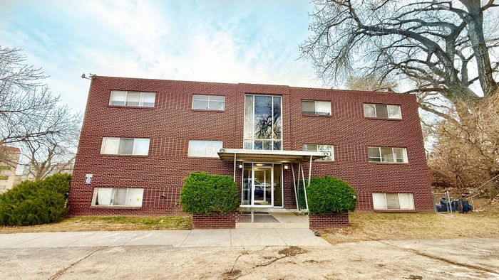 Charming brick apartment building with large windows, surrounded by trees and greenery, highlighting a welcoming entrance.
