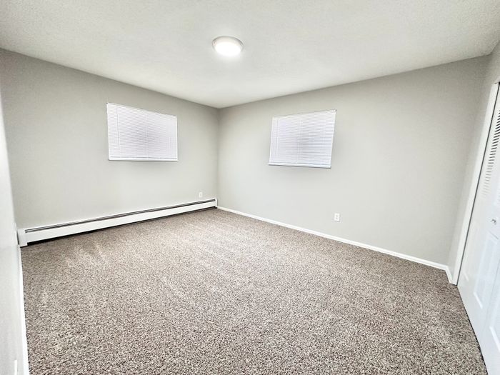 Bright and spacious bedroom with fresh carpeting, neutral walls, and natural light from dual windows.