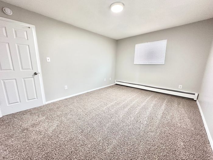 Bright, carpeted bedroom with neutral tones, featuring a closed white door and window blinds for a cozy atmosphere.