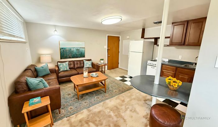 Welcoming living space featuring a cozy brown leather sectional, vibrant teal cushions, and a chic wooden coffee table, seamlessly flowing into a compact kitchen with modern cabinetry and appliances.