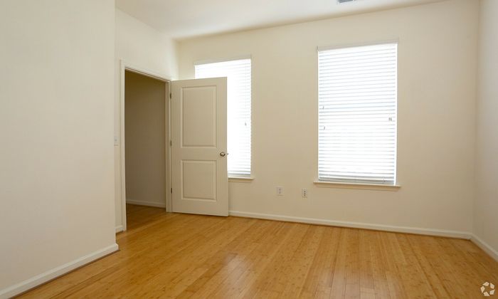 Bright empty room with bamboo flooring, white walls, and two large windows with blinds.