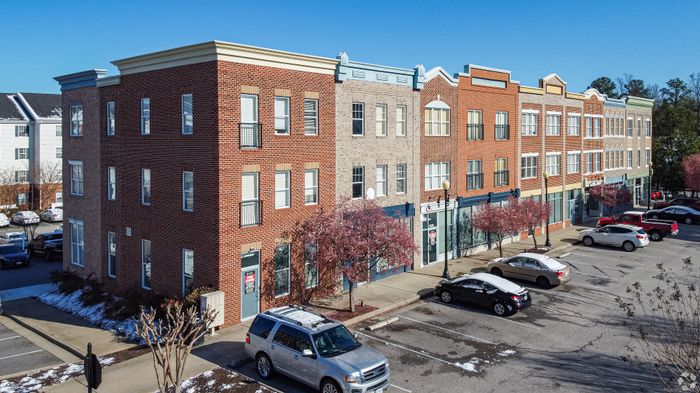 Street view of a modern brick apartment building with retail spaces, surrounded by parked cars and blossoming trees.
