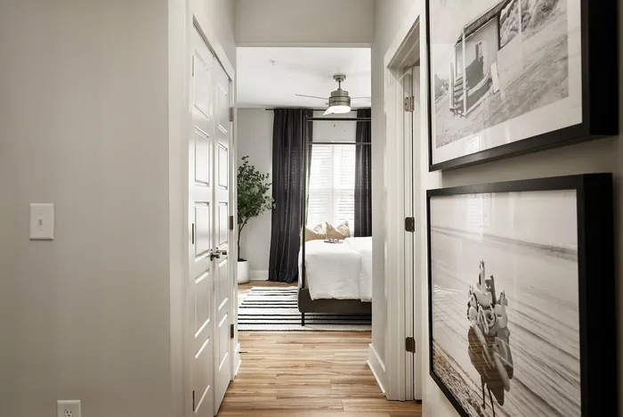 Corridor view leading to a bright bedroom with elegant decor, featuring framed wall art and wooden flooring.
