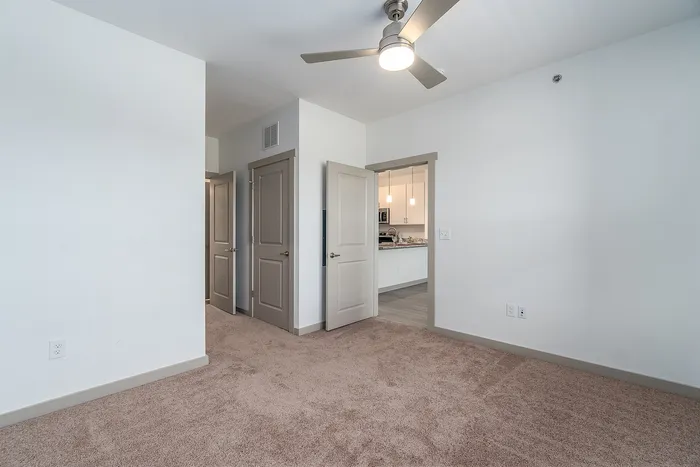 Bright and spacious bedroom with plush carpeting, modern ceiling fan, and an inviting view into the sleek, open kitchen.