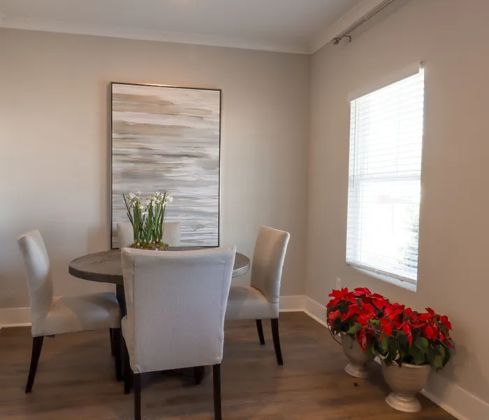 Charming dining area featuring a round wooden table with elegant upholstered chairs, modern artwork, and a window view enhancing natural light, complemented by vibrant potted poinsettias.