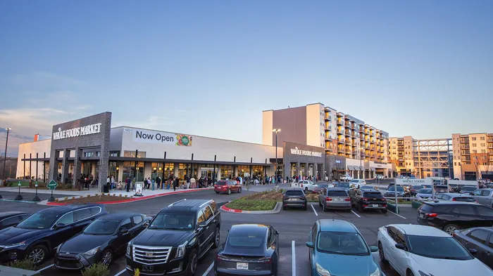 Vibrant urban scene with a bustling Whole Foods Market in the foreground and modern apartments in the background, featuring ample parking and a lively atmosphere.