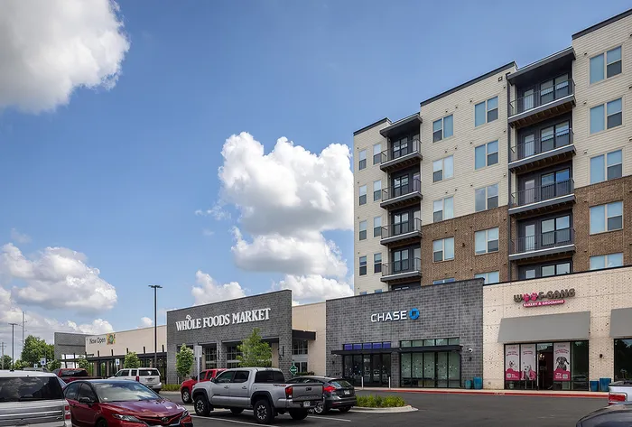Vibrant urban living with modern apartments above retail shops including Whole Foods, Chase Bank, and pet grooming services, set against a backdrop of a clear blue sky.