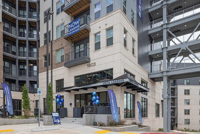 Modern apartment complex exterior with 'Now Leasing' signage, inviting entryway, and vibrant landscaping.