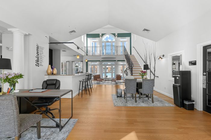 Elegant apartment lobby with a modern open layout, featuring hardwood floors, stylish seating areas, and an inviting staircase with large windows providing ample natural light.