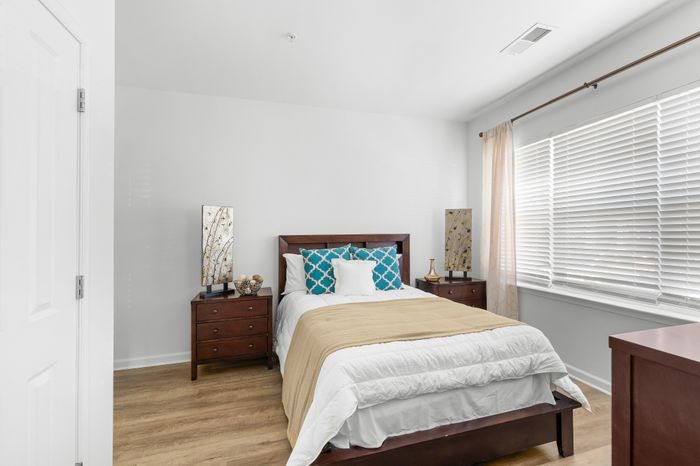 Bright and inviting bedroom featuring a cozy bed with stylish pillows, wooden nightstands, and large window with blinds.