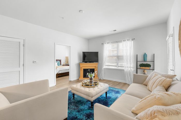 Bright and inviting living room with modern beige sofas, a vibrant blue rug, and a wooden fireplace, leading to a cozy bedroom.