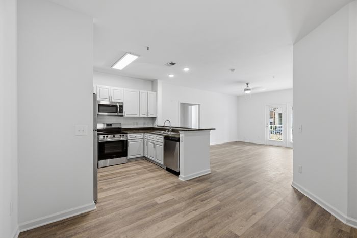 Bright open-concept apartment showcasing a modern kitchen with stainless steel appliances, adjacent to a spacious living area with beautiful wood flooring and ceiling fan, leading to a balcony.
