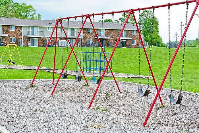 Charming playground with red swings and vibrant green grass, located in a peaceful residential area with brick townhouses in the background.