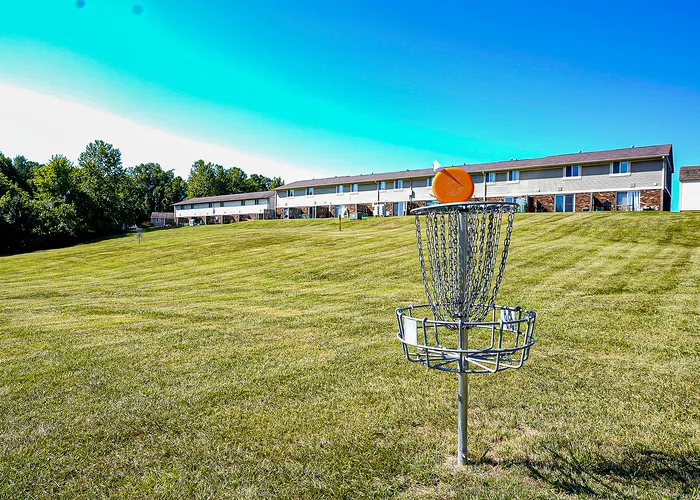 Serene outdoor area featuring a disc golf basket on a lush grassy field, with modern townhouses in the background.