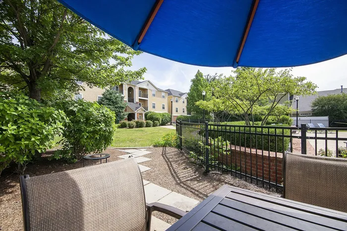 Charming apartment complex with lush greenery and a cozy sitting area under a vibrant blue umbrella. Ideal for relaxation and enjoying outdoor views.