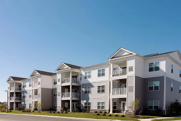 Modern apartment building with three stories, bright white and gray exterior, private balconies, and well-maintained landscaping under a clear blue sky.