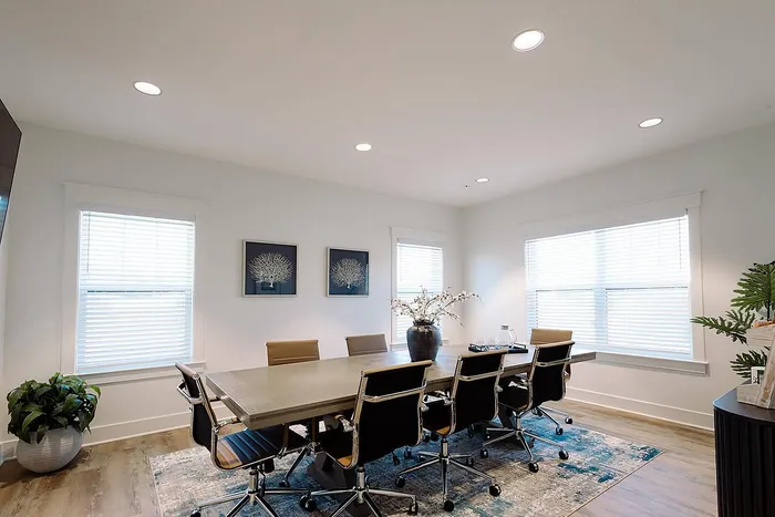 Elegant meeting room with a large wooden table, stylish chairs, and natural light from two windows, complemented by tasteful decor and vibrant greenery.