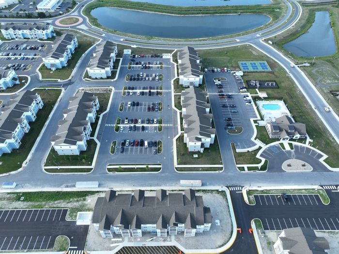 Aerial view of a modern apartment complex with multiple buildings, ample parking spaces, and a scenic pond nearby, offering a vibrant community atmosphere.