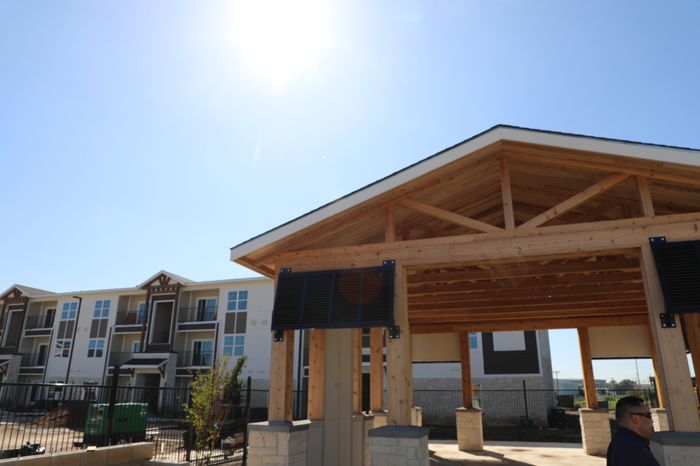 Inviting outdoor view of a modern apartment complex with stylish wooden pergola and sunlit sky, offering a glimpse of upscale living.