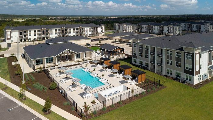 Aerial view of a modern apartment complex courtyard showing a large rectangular swimming pool with striped umbrellas, an adjacent clubhouse, a grilling area, and multi-story apartment buildings under a partly sunny sky.