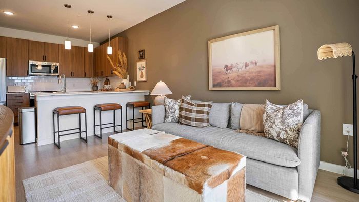 Open floor plan view of a model apartment unit interior, featuring a grey couch with patterned pillows, a framed picture of horses, a cowhide ottoman, a dining area, and a kitchen with wood cabinets and stainless steel appliances.