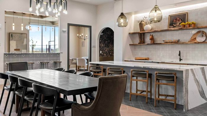 Interior view of an expansive community clubhouse kitchen and dining area, featuring a long dark dining table, modern pendant lighting, leather barstools, and a demonstration kitchen with floating shelves.