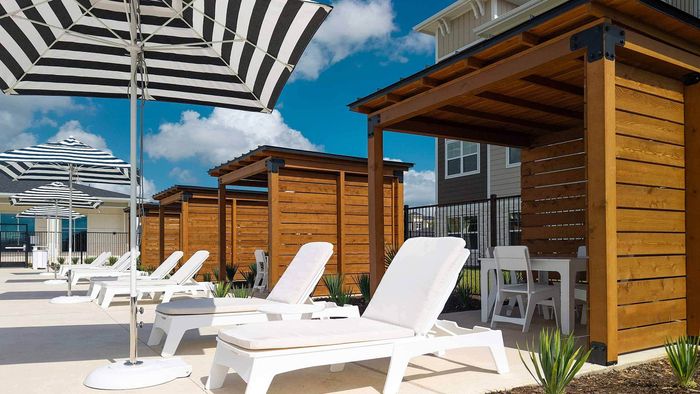 Low-angle view of a sunny pool deck at an apartment complex, featuring white lounge chairs, black and white striped umbrellas, and wooden cabanas with seating areas.