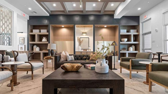Interior view of a sophisticated clubhouse lounge with high, beamed ceilings, a dark chesterfield sofa, grey and green armchairs, built-in shelving, and modern decorative elements.