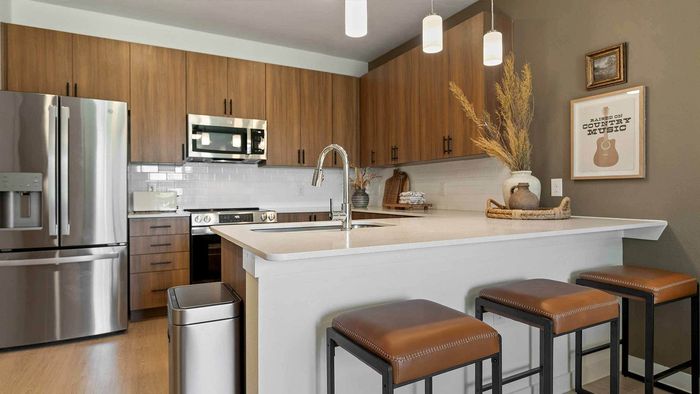 Close-up view of a modern apartment kitchen featuring a white countertop island with an undermount sink, stainless steel faucet, wood cabinetry, a white subway tile backsplash, and stainless steel appliances.