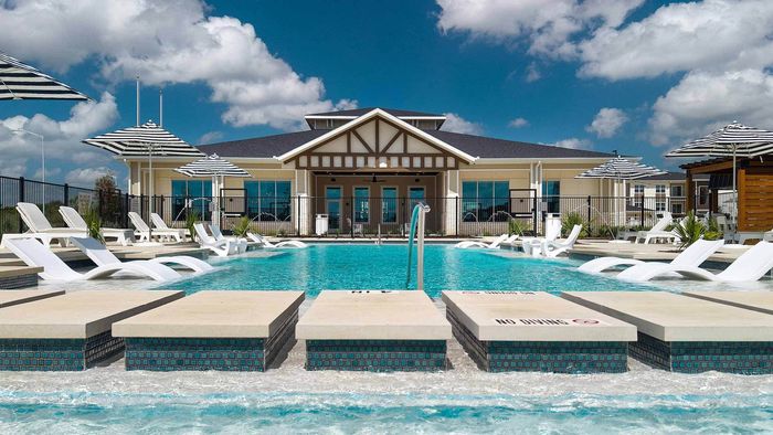 Low-angle view across a resort-style apartment pool from partially submerged lounge chairs on a sun shelf, looking toward the clubhouse building and more umbrellas.