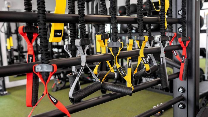 Close-up of modern fitness equipment hanging on a rack in a gym, including yellow and black TRX straps, colorful resistance bands, and a neon 'Let's Do This' sign.