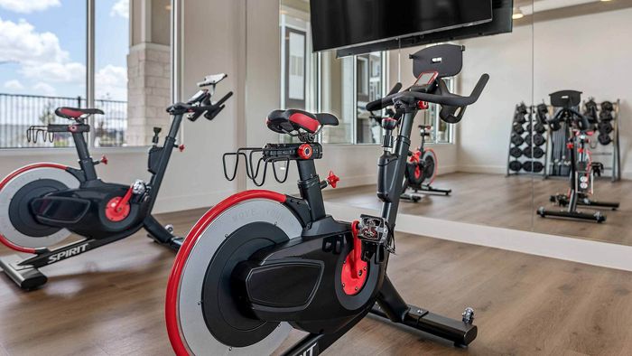 Two modern stationary spin bikes facing a window in a bright apartment fitness center, with a wall of mirrors and a dumbbell rack in the background.