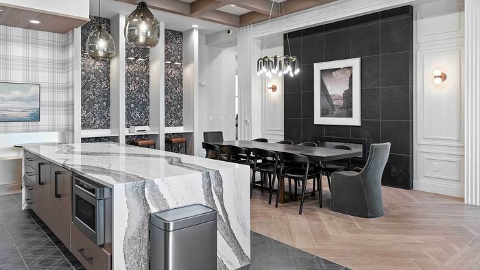 Spacious clubhouse entertaining area featuring a striking waterfall edge quartz island, a long dark dining table with black chairs, unique pendant lighting, and an accent wall with dark floral wallpaper.