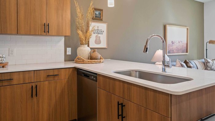 Close-up of a modern apartment kitchen island featuring a stainless steel undermount sink, gooseneck faucet, white quartz countertops, and wood-grain cabinetry.