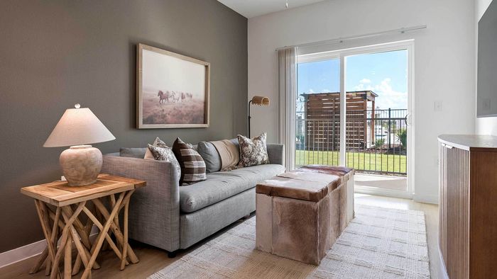 Bright apartment living room with a grey sofa, cowhide ottoman, and sliding glass doors opening to a private outdoor patio area.