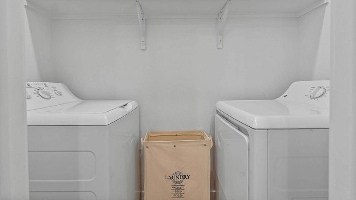 Dedicated apartment laundry closet featuring a side-by-side white washer and dryer set, a canvas laundry basket, and overhead storage shelving.